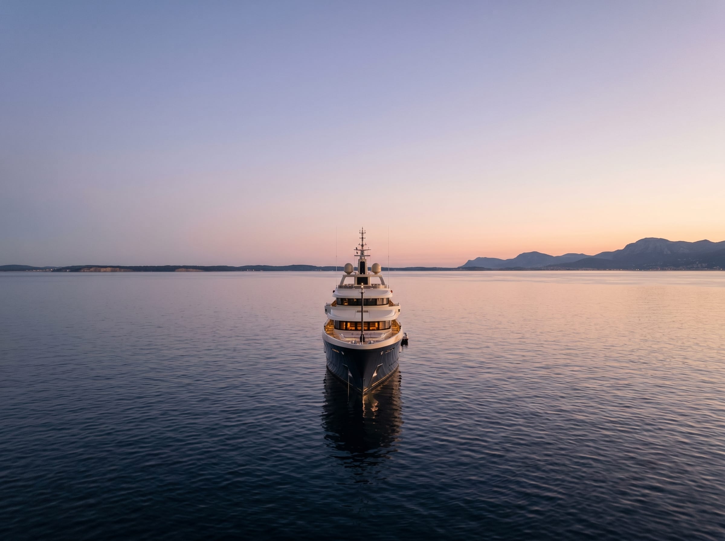 A dark-hulled superyacht on calm open water at dusk, a coastline and mountains behind